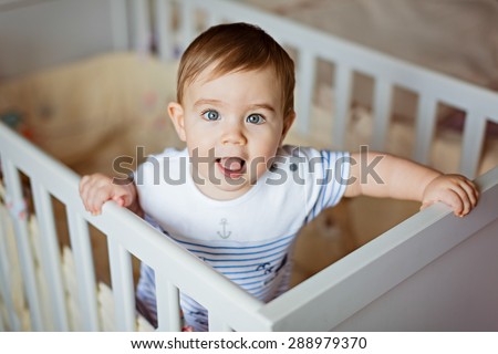 Little cute adorable little blond boy in a striped bodykit is in the nursery with white crib and laughs