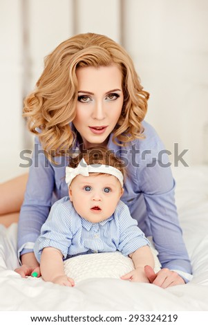 Portrait of a beautiful mom blonde and a little girl baby with blue eyes in a striped blue dress lying on white bed.