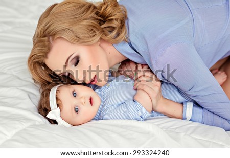 Portrait of a beautiful mom blonde and a little girl baby with blue eyes in a striped blue dress lying on white bed. Mommy holds his daughter\'s hands and kisses her