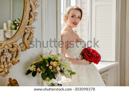 Charming beautiful young blonde girl with red lipstick on my lips, with red roses and white lace dress standing at the window in the interiors of the house