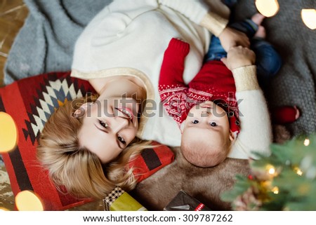 Charming toddler boy red sweater and his mother\'s blonde, lying on the floor on cushions on the Christmas tree and garlands in the house and smile