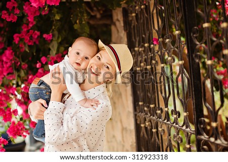 Beautiful young mother in a hat and white blouse holding a small son baby with big brown eyes and laughing merrily, summer amid flowering Bougainvillea pink flowers