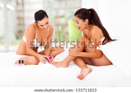 two happy lady friends painting toe polish on bed at home