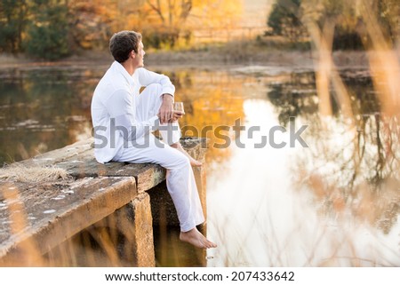 young man sitting on pier with wine and enjoying sunset view by the lake