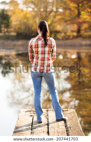 back view of peaceful young woman standing on lake pier in fall