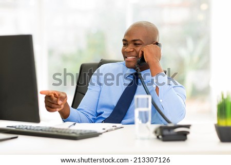 young african businessman talking on landline phone and pointing at computer screen