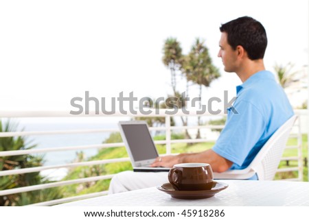 casual businessman using laptop on balcony with sea view behind, shallow focus on the coffee cup