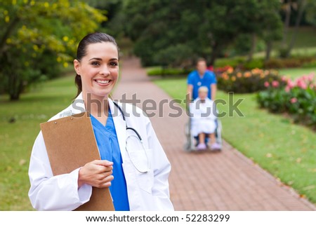 friendly female doctor portrait outdoors, background is her colleague pushing patient in wheelchair