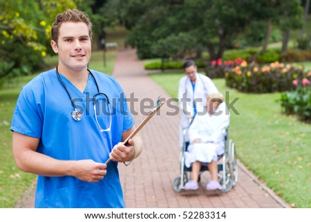friendly young male doctor portrait outdoors, background is his colleague pushing recovery, patient in wheelchair