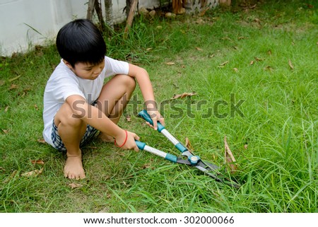 Young asian boy use hand grass scissors to cut the long grass at home garden with tired and boring face.