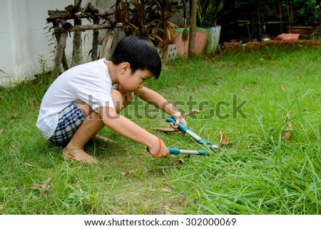 Young asian boy use hand grass scissors to cut the long grass at home garden with tired and boring face.