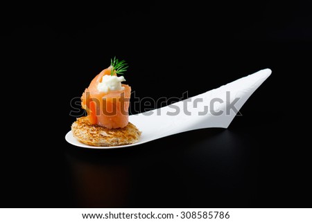 Mini snacks with smoked salmon, cheese, fresh dill and toast bread on a white, designer spoon, with a black background.