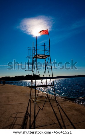 Lifeguard tower on a beach with red flag marking closed beach and forbiden swimming. The sun is shining thru the flag on top of a baywatch tower and silhuettes of couple walking are visible.