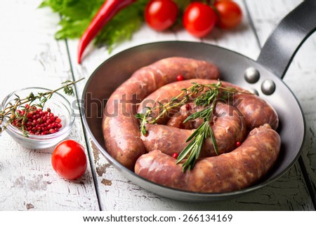 Raw sausages in a frying pan on a wooden white background. Vegetables, green salad, spices and herbs