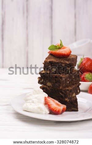 Stack of square pieces of chocolate brownies with hazelnuts, served with whipped cream, strawberry and mint on white wooden table. Delicious breakfast concept. Space for text. Selective focus