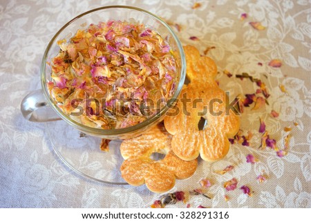 Gentle composition of the drink of tea rose petals, transparent cup and saucer, cookies in the shape of flowers and scattered petals