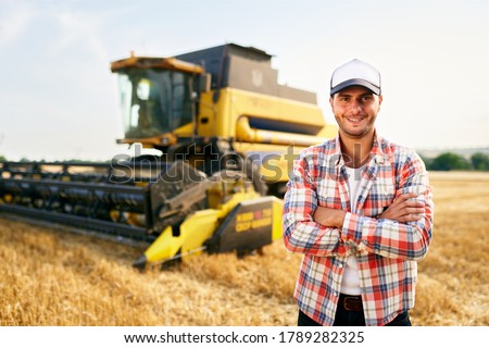 Portrait of proud harvester machine driver with hands crossed on chest. Farmer standing at his combine. Agronomist looking at camera. Rancher at harvesting work on stubble of harvested wheat field.
