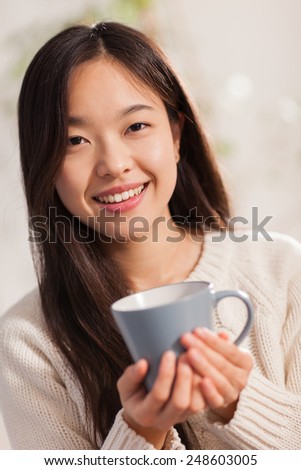 photo of young asian woman with hot mug in her hands smiling towards the camera