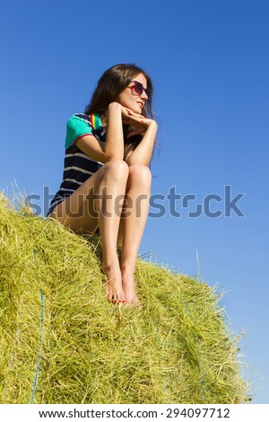 Pretty lady is sitting on the big haystack and looking into the distance.