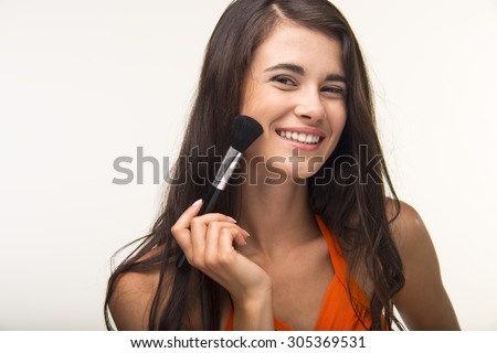 The brunette with long hair is holding makeup brush. Good-looking girl in orange shirt is smiling.