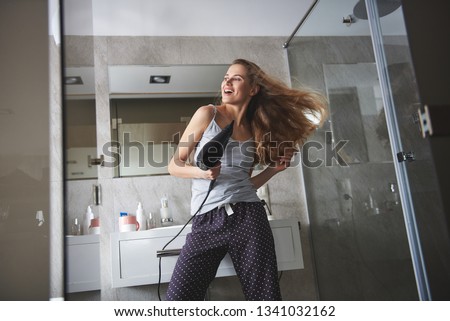 Happy in ordinary thing. Waist up low angle portrait of happy smiling woman having fun and dancing while drying her voluminous hair by hairdryer in bathroom 商業照片 © 