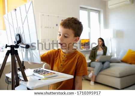 Cute little boy painting picture on easel while mother using laptop stock photo