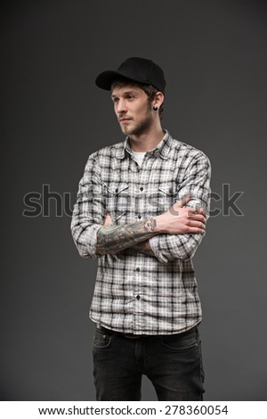 guy holds his hands together on his chest. dressed in a plaid shirt and a black cap. his body has tattoos and piercings in the ears