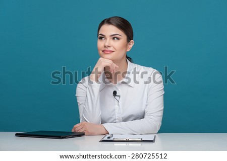 Waist up portrait of elegant woman reporter with Caucasian appearance, who is smiling and dreaming about something while sitting at the table and propping up her head with her right hand.