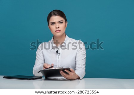 Waist up portrait of elegant woman reporter of Caucasian appearance, who is looking at the camera questioningly and trying to understand something holding the folder and pen in her arms.