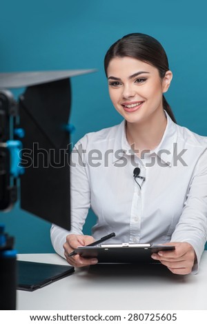 Waist up portrait of elegant woman reporter, who is smiling and looking at the camera, which is visual on the foreground, while sitting at the table and holding the folder and pen.