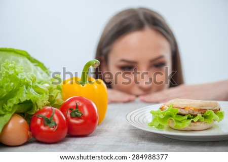 Beautiful young girl is looking at the hamburger with appetite. She makes her decision to eat it and does not look at vegetables. Focus on hamburger and vegetables. Isolated on a white background