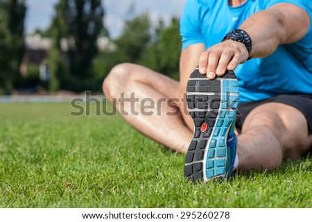Close up of legs and torso of fit sportsman sitting on grass. He is stretching his left hand and doing warm-up.  There is copy space in left side