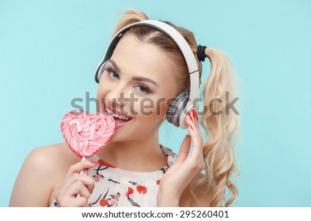 Beautiful girl is eating lollipop in shape of heart. She is listening to music from headphones. The lady is smiling with enjoyment. Isolated on blue background and there is copy space in right side