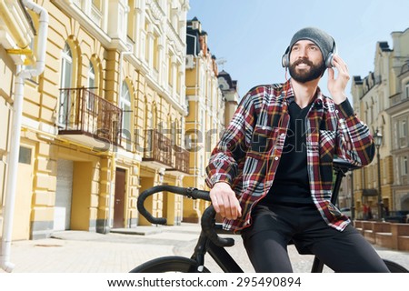 Handsome young hipster guy is sitting on bicycle. He is listening to music with enjoyment. The guy is smiling and looking aside
