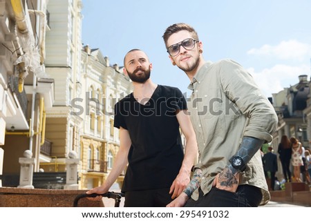 Handsome two friends are standing and looking interestingly straight. One man is holding bicycle. Another man with glasses keeps his hand in his pocket