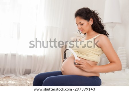 Cute young pregnant woman is touching headphones to her large abdomen. She lets her baby to listen to music. The woman is smiling gently. Copy space in left side