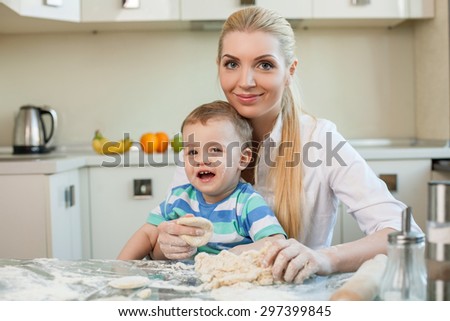 Cheerful mom and her small son are baking cookies. The woman is holding toddler on her knees. They are smiling and looking at the camera with joy. Copy space in left side