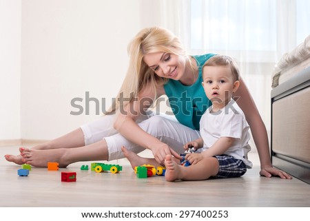 Attractive mother plays with her child on the floor. She is smiling and looking at toys. Her son is looking at the camera seriously
