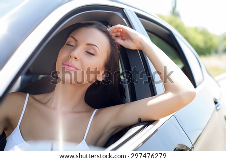 Cheerful girl is driving her new car with pleasure. She is closing her eyes with enjoyment and smiling. The lady raises her elbow through the window and touching her hair gently