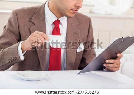 Close up of body of man in suit holding a folder and reading documents. He is sitting at the table in restaurant and drinking coffee