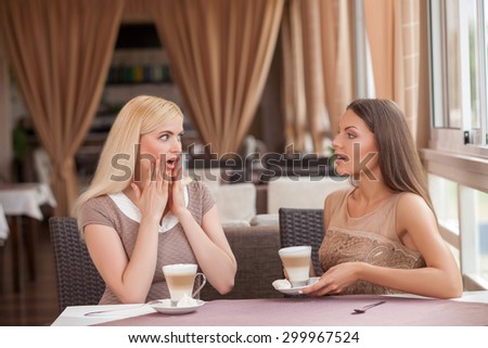 Attractive woman are talking in cafe. One woman is telling s story to her friend with seriousness. Another girl is shocked. They are drinking latte and sitting at the table