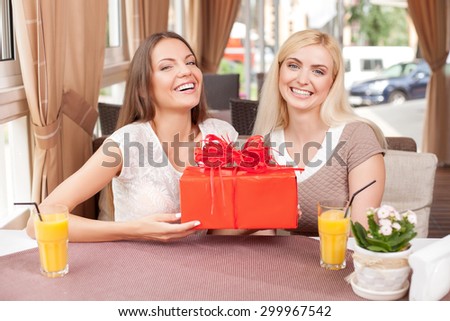 Cheerful women are giving a gift to each other in case of celebration. The brunette is holding a box of gift in her hands happily. The friends are smiling