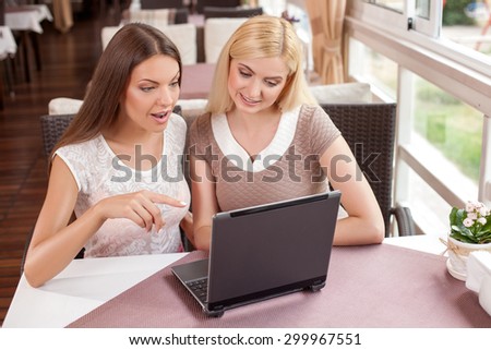 Pretty girls are sitting at the table in cafeteria. They are looking at a notebook with interest and smiling. The brunette girl is pointing her finger at it with shock