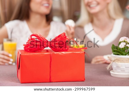 Close up of red box of a gift on a table. Two women are sitting behind it and smiling. They are eating sweet food and drinking juice