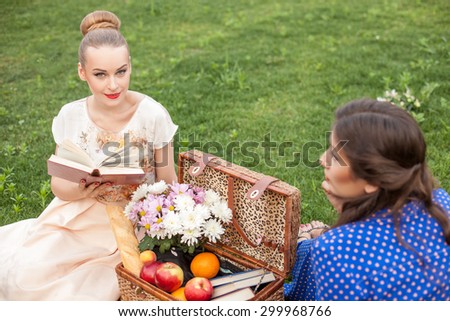 Beautiful women are resting and sitting on grass near the basket of food. The blond girl is reading a poem aloud and smiling with pleasure. Her friend is listening to her attentively