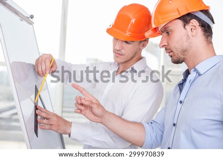 Attractive architects are working on a project. One builder is drawing sketches on blueprint with inspiration. Another man is pointing his finger at it and giving advices concerning a plan seriously
