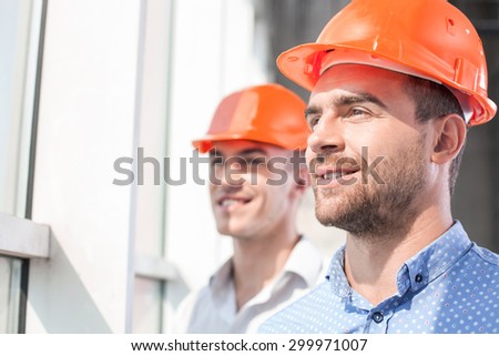 Handsome foreman and architect are standing and looking through the window dreamingly. They are smiling with hope. Their project was approved