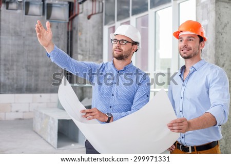 Attractive architect is explaining to foreman the plan of building. He is pointing his hand aside. The men are holding a paper of sketches and smiling dreamingly