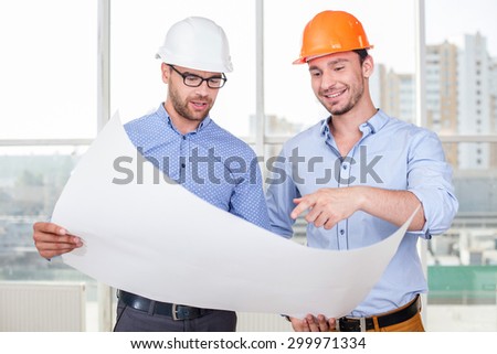 Handsome architect is showing to a foreman the plan of a building with seriousness. The foreman is looking and pointing his index finger at it. He is smiling