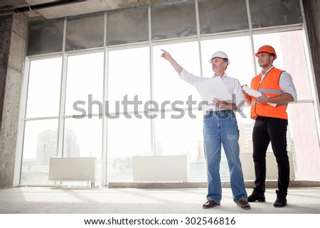 Senior architect and young builder are discussing new project. The architect is pointing his finger sideways seriously. The foreman is looking there with interest and smiling. Copy space in left side